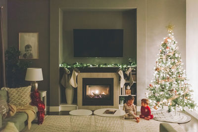 Cute kids sitting by illuminated christmas tree at home