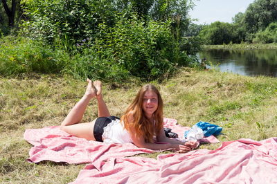 Red-haired girl sunbathes on a pink bedspread on the riverbank on a sunny summer day.
