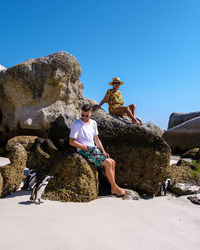 Rear view of man sitting on rock at beach against clear blue sky