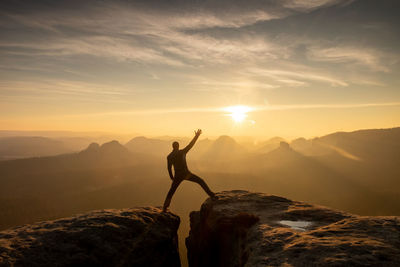 Young man standing on a cliff edge celebrates reaching the top of  mountain, arms in air, shouting.