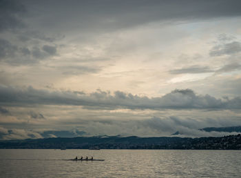 Distant view of people sculling in lake against cloudy sky