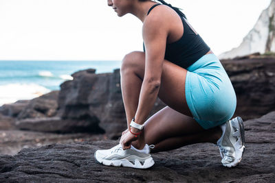 Side view of fit ethnic female athlete tying shoelaces on sneakers while sitting on seashore and preparing for workout