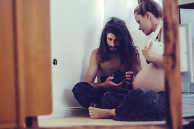 Pregnant woman sitting by shirtless man on floor at home