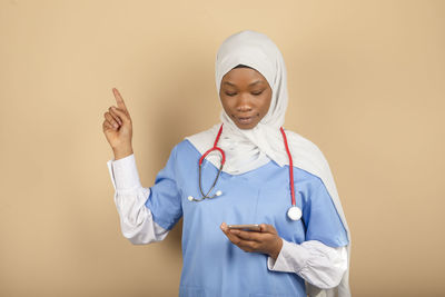 Online nurse with headscarf isolated on yellow background.holds smartphone, points finger up person