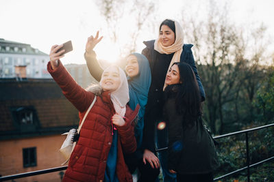 Happy young muslim woman taking selfie with friends by railing in city