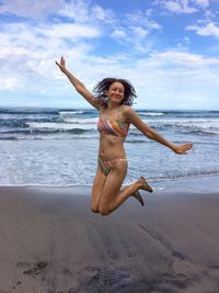 Full length of woman in bikini jumping at beach against sky
