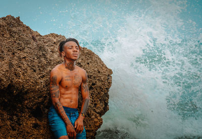 Portrait of shirtless man standing on rock at beach
