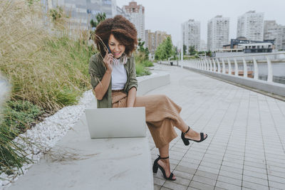 Young businesswoman talking on mobile phone and using laptop sitting at promenade