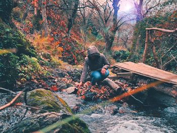 Woman wearing hood crouching at riverbank in forest during autumn