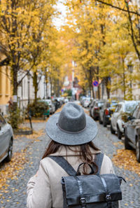 Rear view of young woman wearing backpack, walking in city street with autumn trees