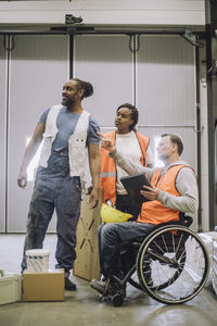 Carpenter sitting on wheelchair with digital tablet instructing male and female coworkers in warehouse