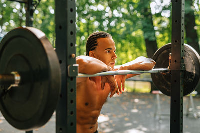Shirtless young man standing by barbell against tree