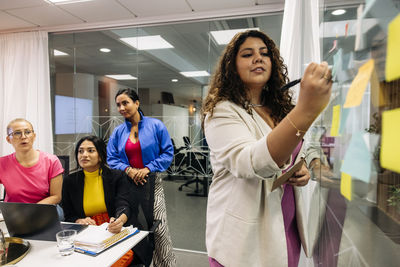 Female entrepreneur explaining colleagues while writing on glass wall at office