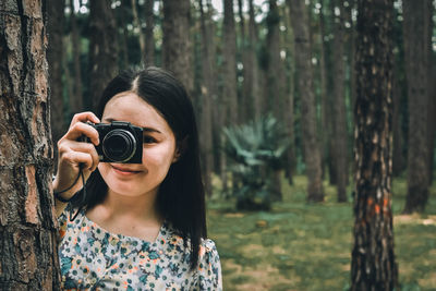 Portrait of woman photographing through tree trunk in forest