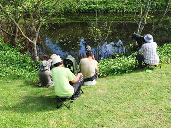 Rear view of people photographing by lake on grassy field during sunny day at park