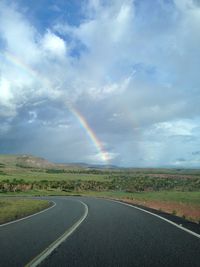 Rainbow over road amidst field against sky