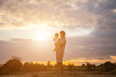 Father carrying daughter while standing on land against sky during sunset