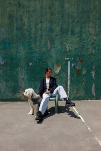 Full body of fashionable young ethnic female with dark hair in stylish outfit sitting on stool near adorable fluffy old english sheepdog near weathered wall on sunny day