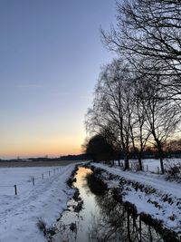 Bare trees on snow covered field against sky during sunset