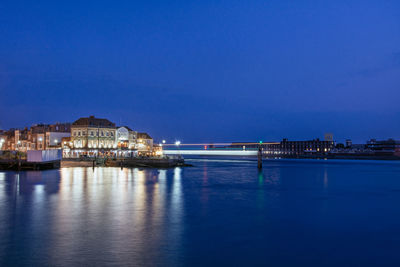 Illuminated buildings by sea against blue sky at dusk