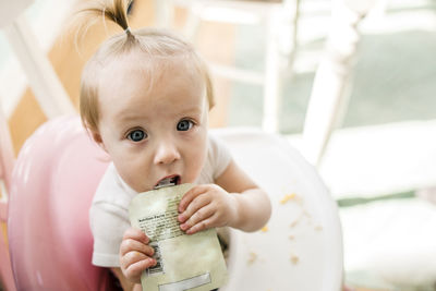 High angle portrait of cute baby girl eating apple sauce from pack while sitting on high chair at home