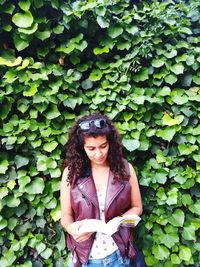 Woman reading book while standing against plants