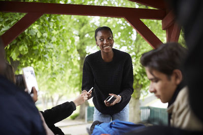 Smiling female teenager looking at friend while collecting smart phone at park