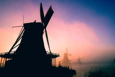 Silhouette traditional windmill by sea against sky at sunset