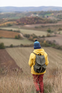 Rear view of a small boy with a backpack standing on a hill in autumn day and looking at view