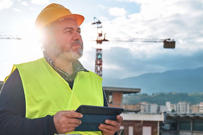Portrait of middle aged bearded supervisor in hardhat and safety vest with tablet on building site.