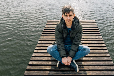 An adult man sits cross-legged on a wooden pier on a lake in jeans and a jacket