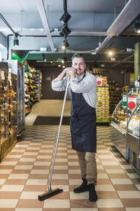 Full length portrait of smiling mature entrepreneur holding broom while standing in store