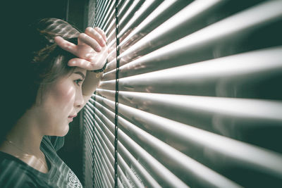 Side view of thoughtful woman looking through window blinds