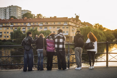 Rear view of male and female friends standing near railing at sunset