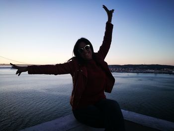 Woman sitting on railing by sea against clear sky during sunset