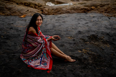 Portrait of smiling young woman with shawl sitting on black sand beach