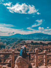 Man sitting on railing by mountain against sky