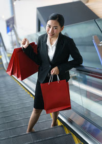 High angle portrait of woman holding shopping bags while standing on escalator