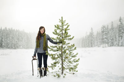 Portrait of woman holding pine tree while standing on snow covered field against sky in forest