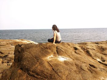 Rear view of young woman sitting on rock at shore against clear sky