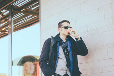 Fashionable man drinking coffee against wall outdoors