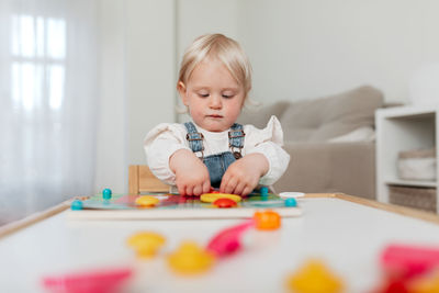 Charming little kid with blond hair assembling game pieces at table at home