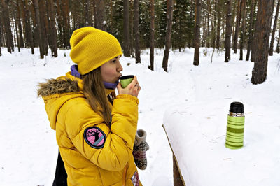 Young woman in yellow jacket drinking hot tea from green thermos on a hike in a winter snowy forest