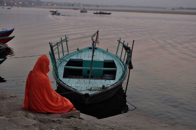 Man covered in orange fabric sitting by moored boat in ganges river during sunset