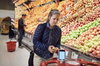 Woman holding apple by shopping cart against man standing at supermarket