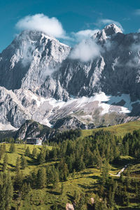 Day photo of a rocky mountain partly covered with snow, in the clouds and a green forest below.