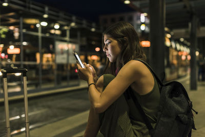 Young woman with backpack and baggage waiting at the station by night using cell phone