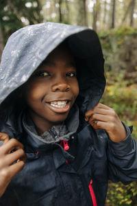 Portrait of boy with toothy smile holding hood of raincoat in forest