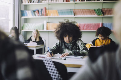 Boy with afro hairstyle writing in book while sitting at desk in classroom