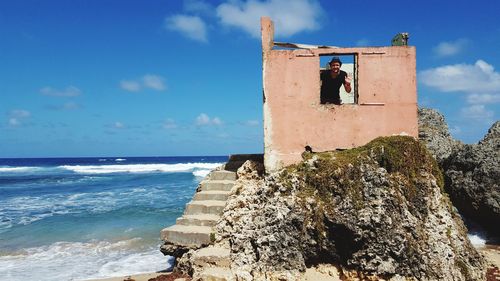 Man waving through window of built structure on rock by sea against blue sky